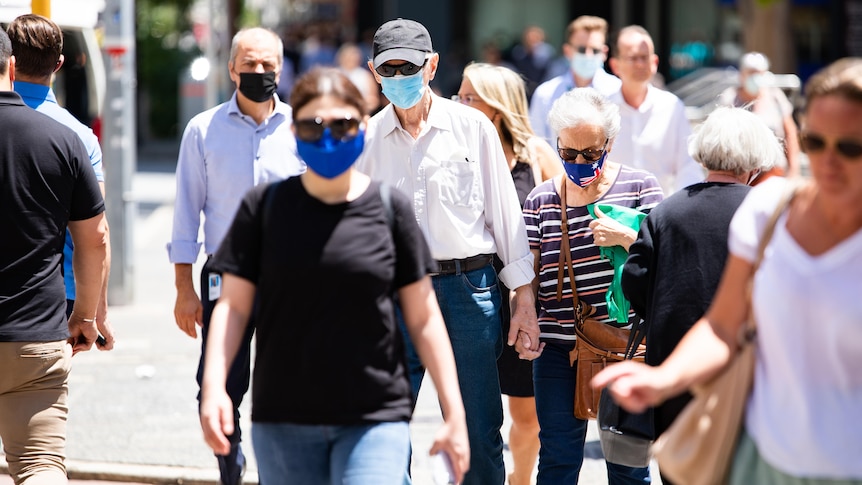 A crowd of people walk across the street in masks, including an older couple.
